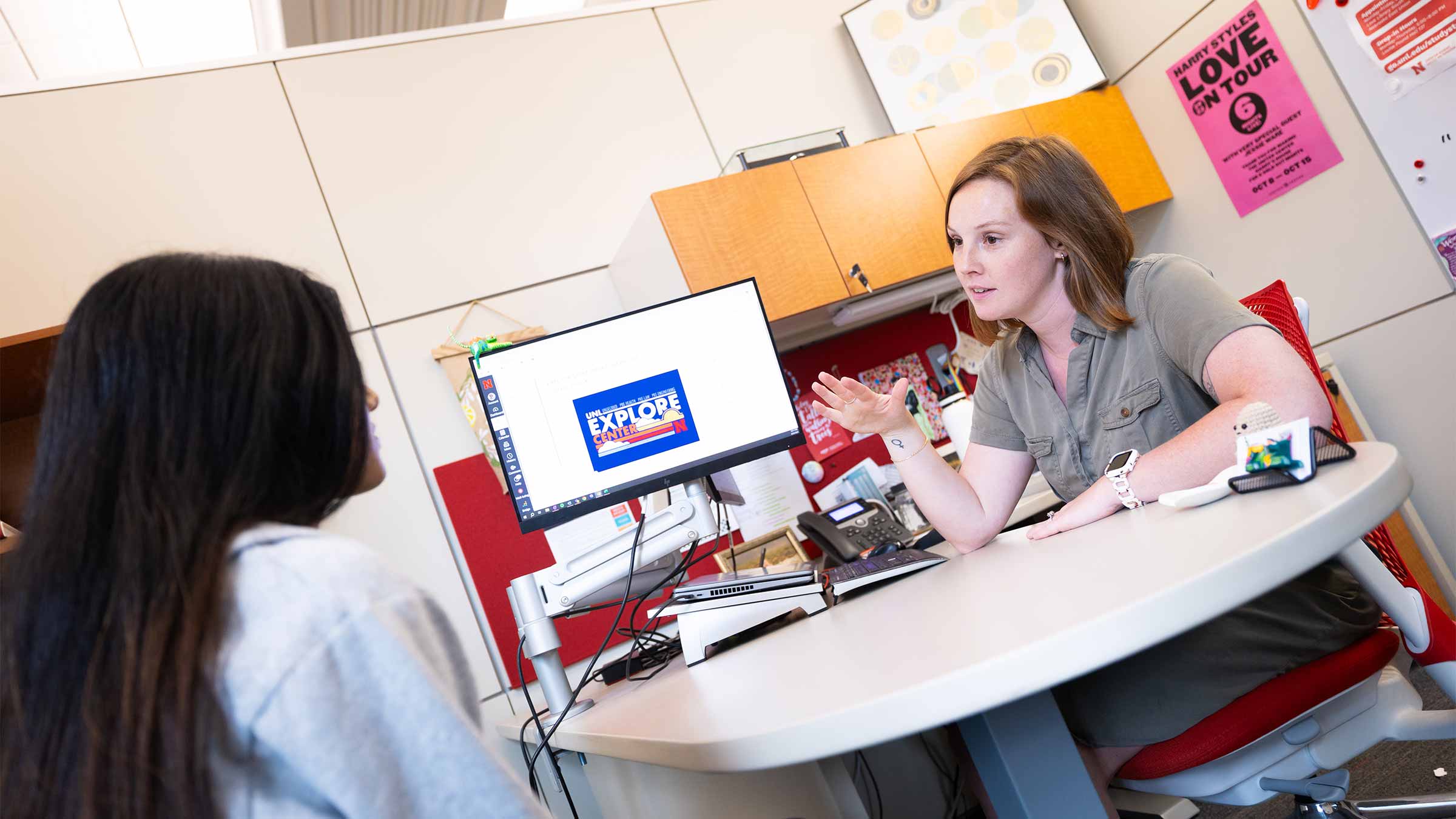Academic Navigator and student talking at a desk in an office with a computer and colorful posters on the wall.