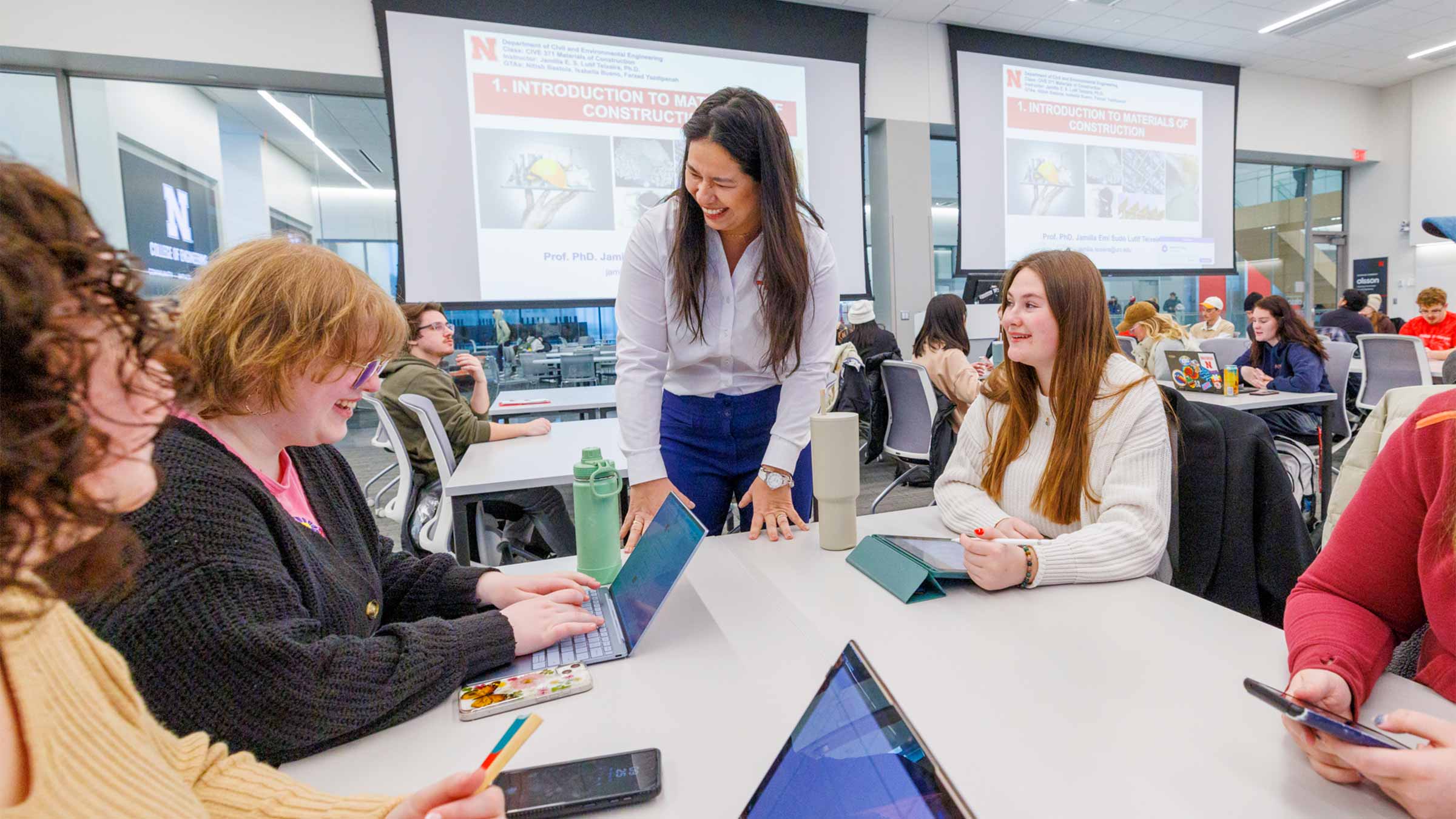 Students interact with a professor in a classroom, using laptops and tablets.