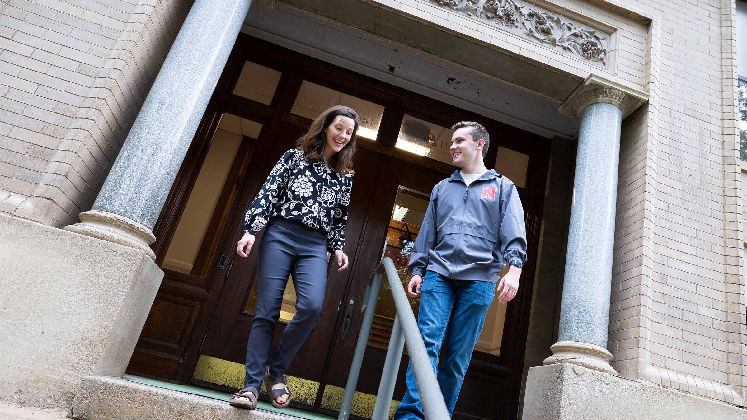 Two people walking down steps of a building with large wooden doors and stone columns.